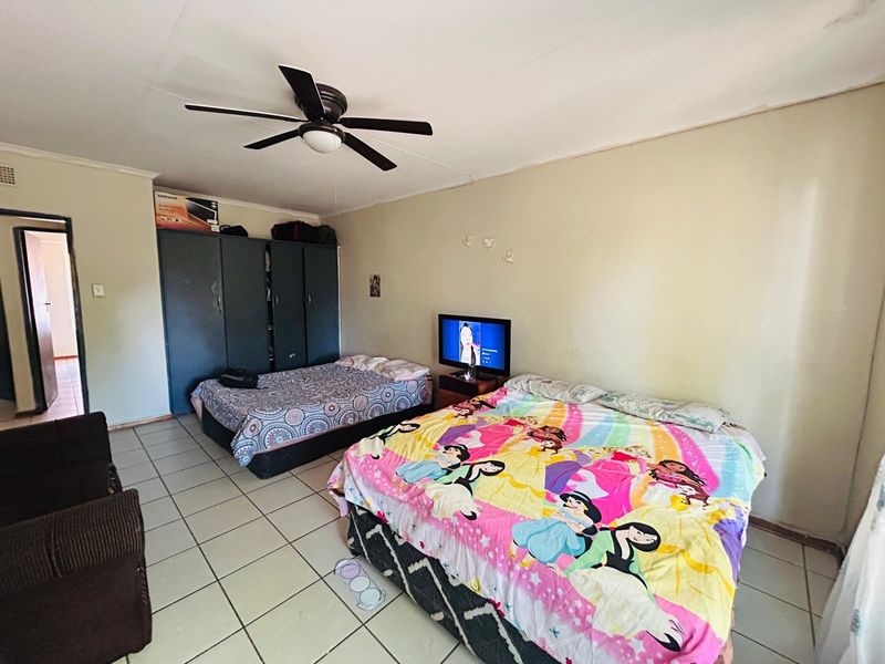A spacious bedroom featuring tiled flooring, a ceiling fan, and built-in cupboards, offering a functional layout with potential for cosmetic updates.