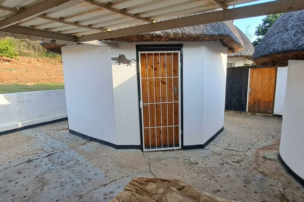 A view of a thatched-roof structure with a covered patio area and concrete paving, featuring a secure timber entrance door.