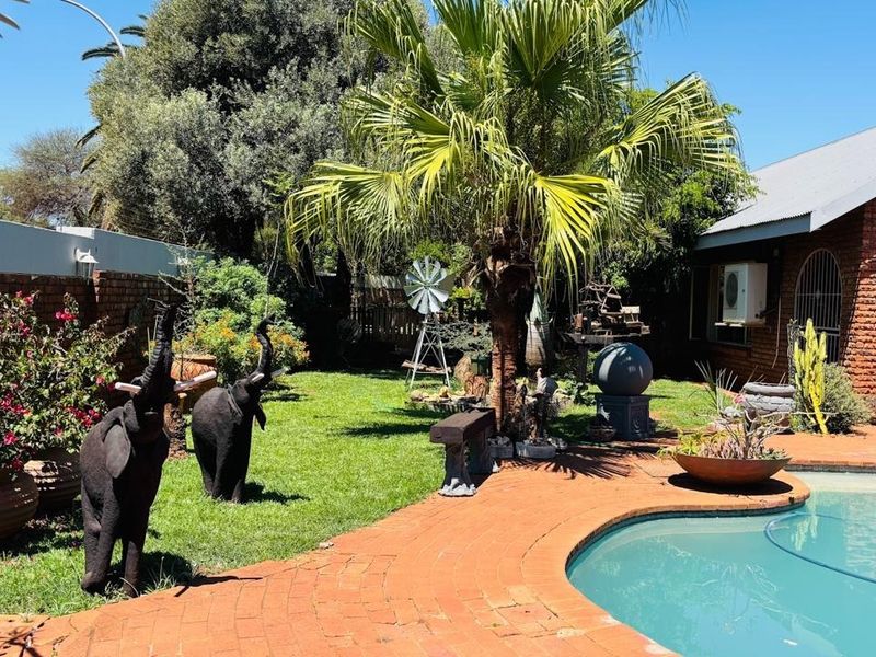 A well-maintained outdoor pool area featuring a curved brick patio, established garden landscaping, and a brick-faced house exterior with an arched entrance.