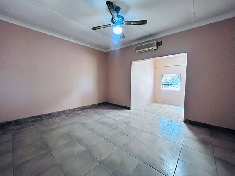 An empty living area featuring tiled flooring, a ceiling fan, and a wall-mounted air conditioning unit, leading into an adjacent secondary space.
