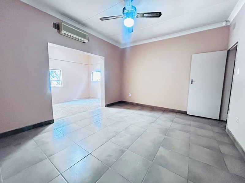 An empty living area featuring tiled flooring, a ceiling fan, and a wall-mounted air conditioning unit, with an adjoining room visible through the opening.
