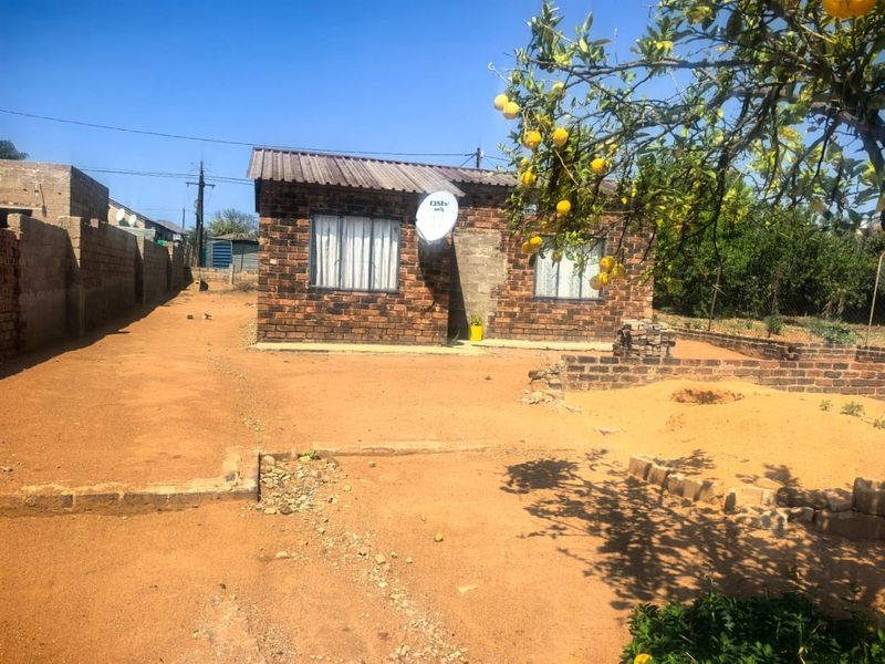 A single-story face brick house featuring a corrugated metal roof and a satellite dish, situated on a large plot with an unpaved yard.