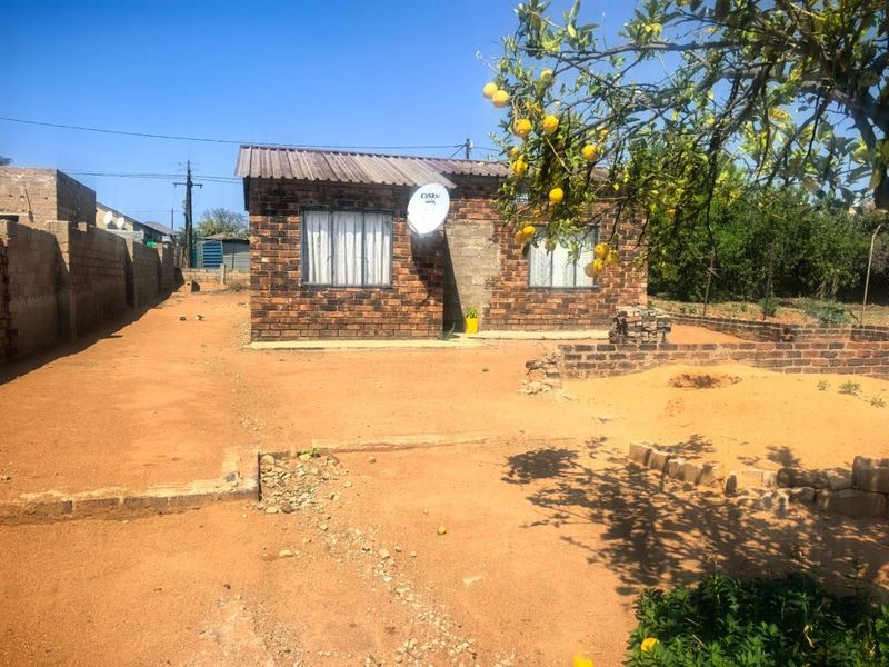 A single-story face brick house featuring a corrugated metal roof and satellite dish, situated on a large, unpaved plot with perimeter walling.