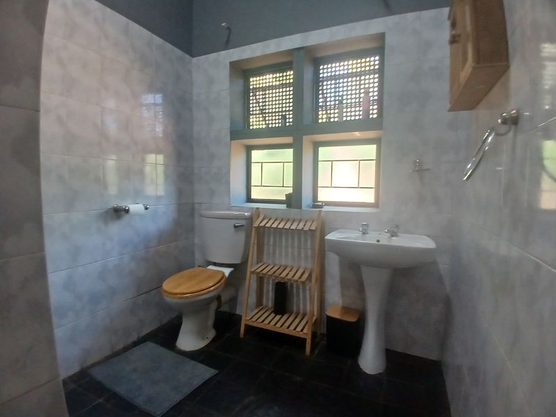 A functional bathroom featuring a pedestal washbasin, toilet, and tiled finishes with security-barred windows.