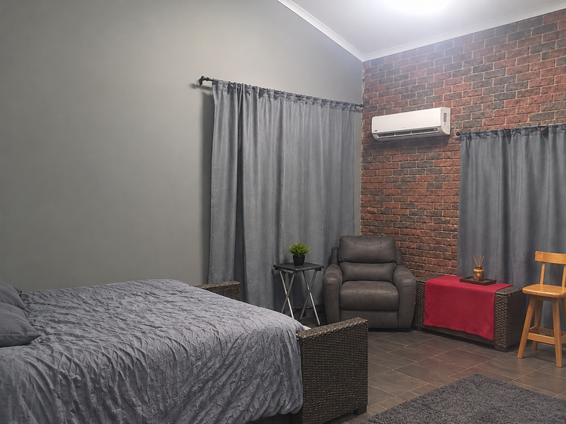 A spacious bedroom featuring a vaulted ceiling, exposed brick feature wall, tiled flooring, and a wall-mounted split system air conditioner.