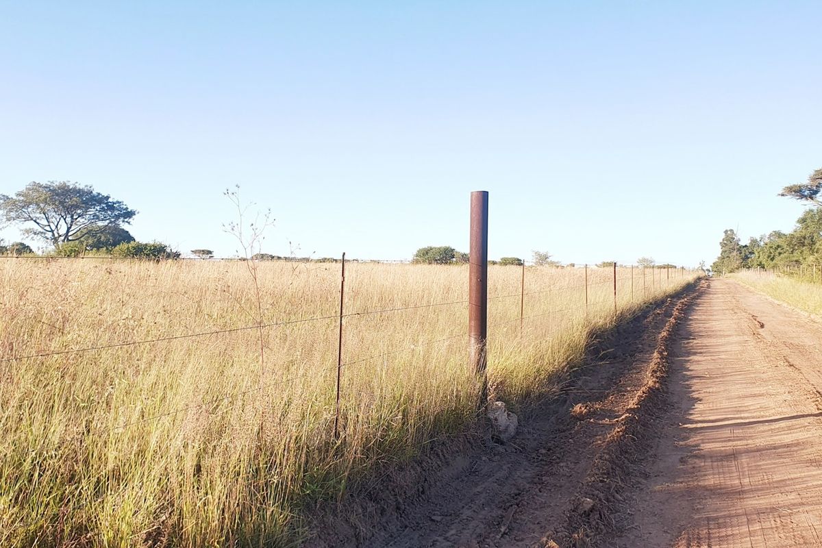 A view of a dirt road bordering a vacant residential plot enclosed by a wire fence with metal posts.