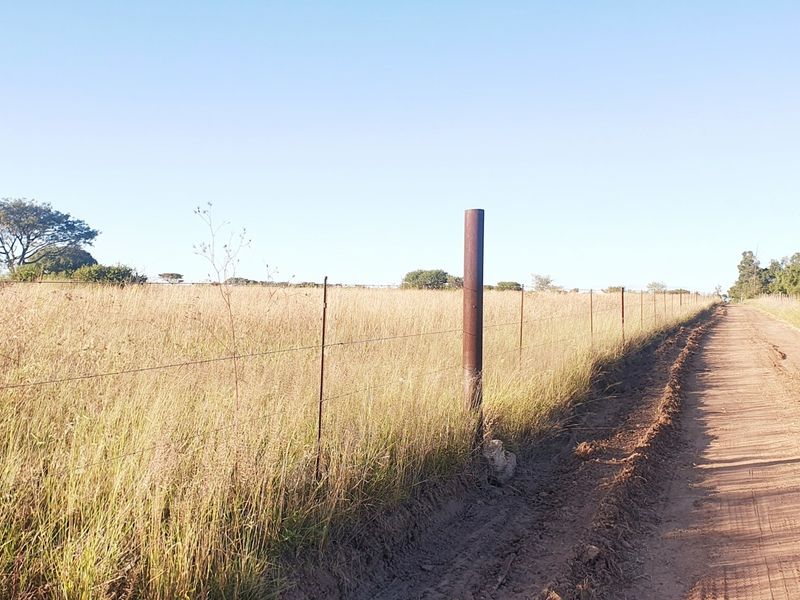 A view of a dirt road bordering a vacant residential plot enclosed by a wire fence with metal posts.