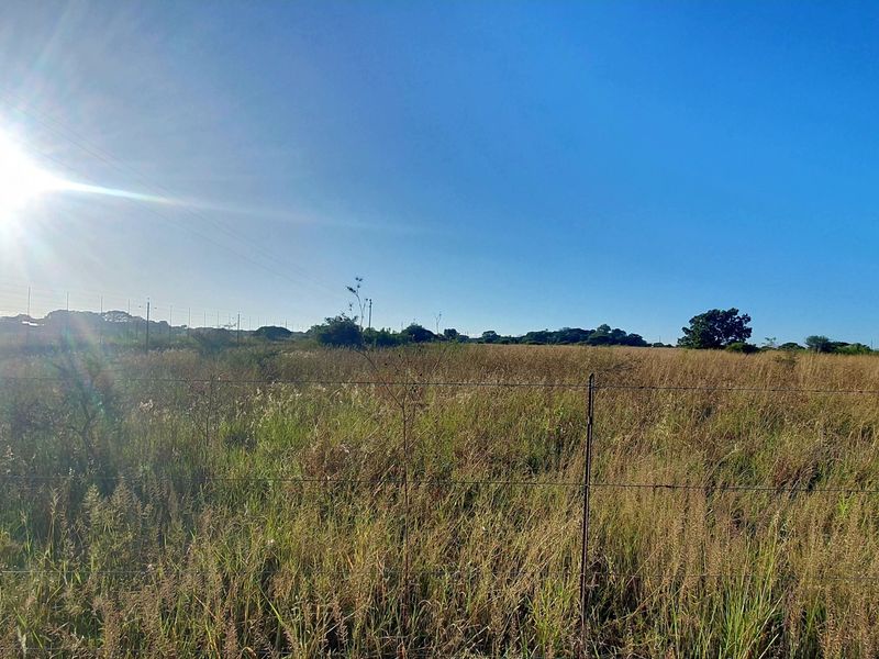 A wide-angle view of a vacant residential plot featuring natural grass cover, perimeter wire fencing, and utility infrastructure under a clear sky.