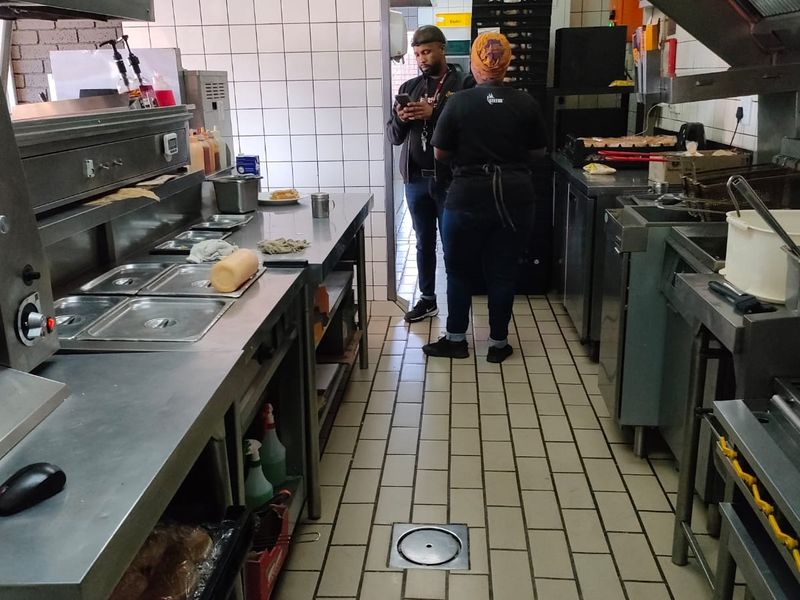 A functional commercial kitchen space featuring stainless steel work surfaces, industrial-grade deep fryers, an extractor hood, and durable tiled flooring with integrated drainage.