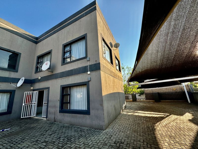 Exterior view of a multi-story apartment building featuring a paved driveway, shade cloth carport, and secure gated entrance.