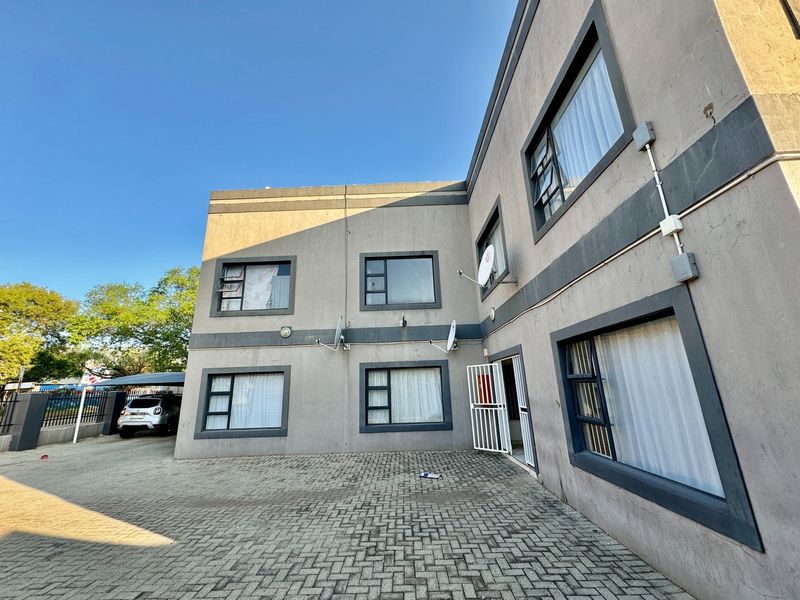 Exterior view of a multi-story apartment building featuring paved driveway access, aluminium window frames, and security entrance gate.