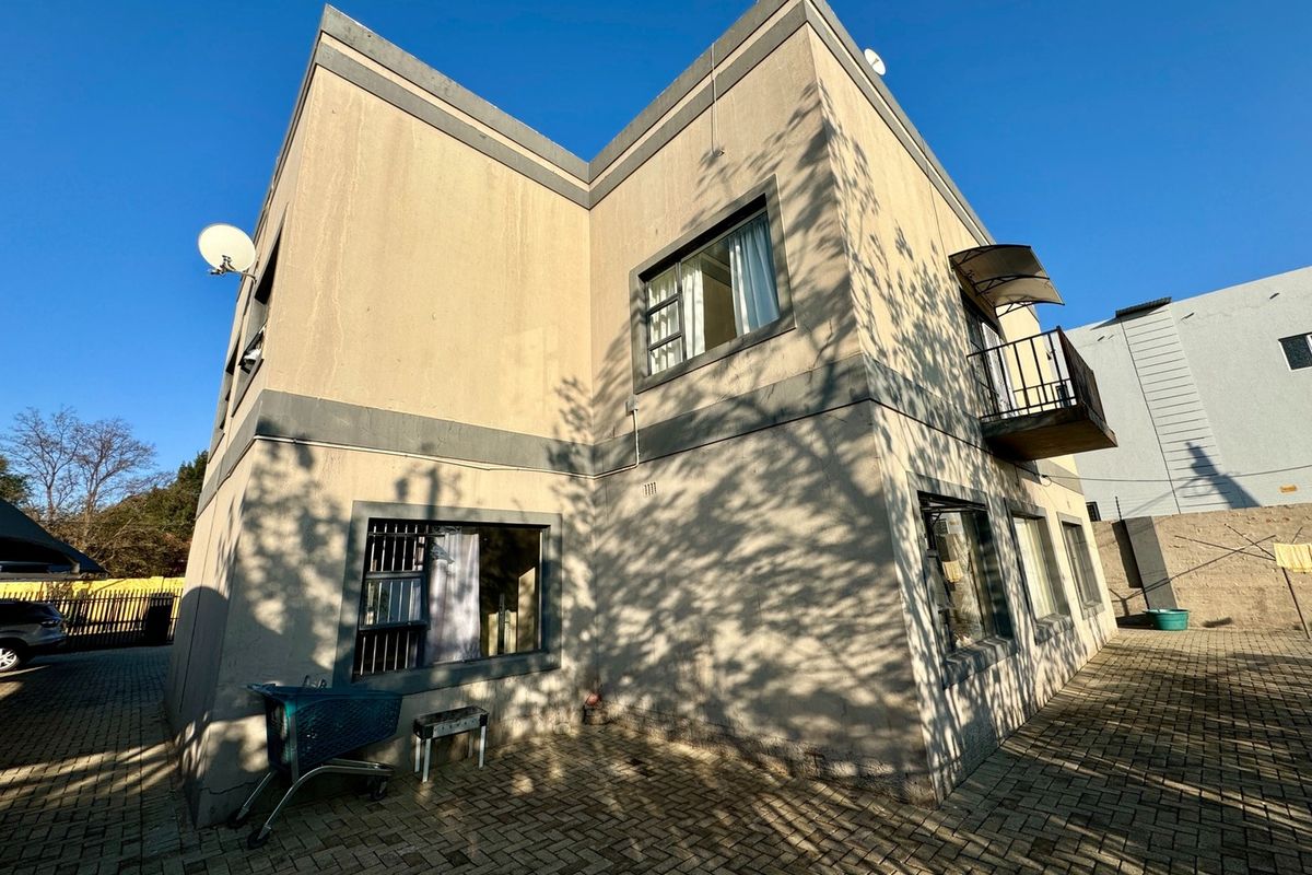 Rear exterior view of the apartment building featuring a paved courtyard, balcony with metal railing, and security bars on ground floor windows.