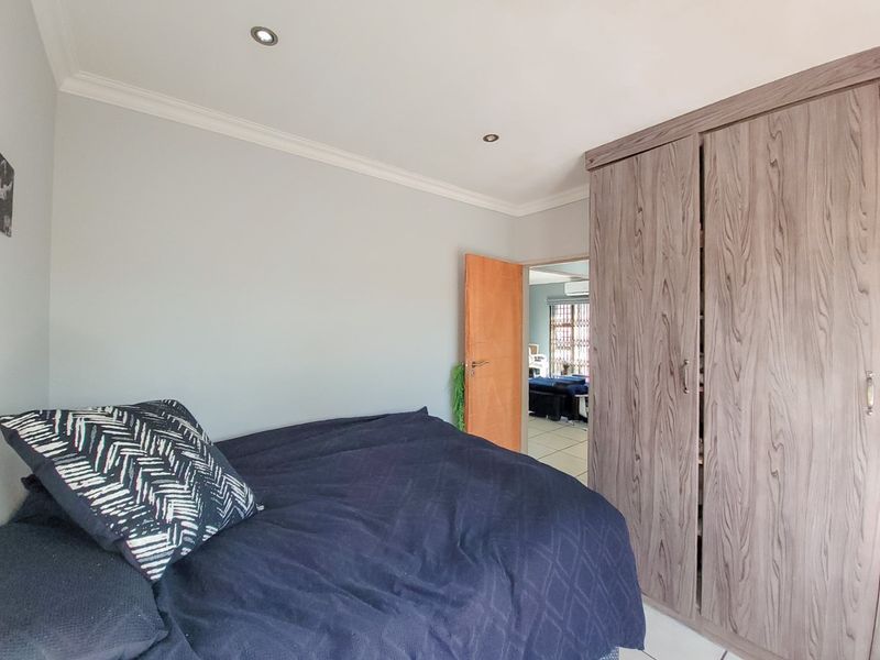 A bedroom featuring built-in wood-grain wardrobes, recessed downlighting, and neutral wall finishes, with an open doorway leading to an adjacent living area.