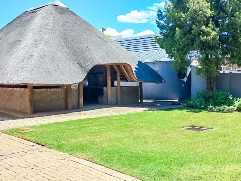 A spacious rear garden area featuring a large thatched lapa with a built-in braai, brick paving, and a well-maintained lawn.