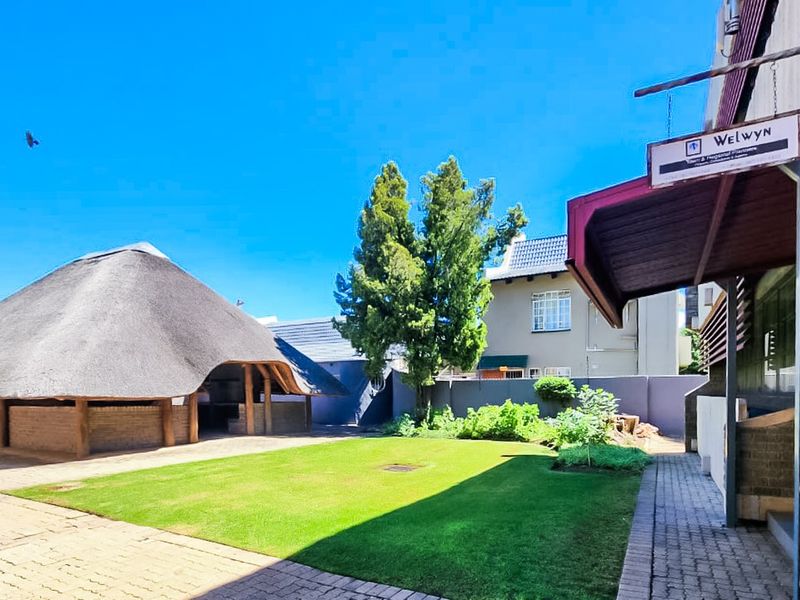 A well-maintained commercial courtyard featuring a large thatch-roofed lapa, paved walkways, and a central lawn area.