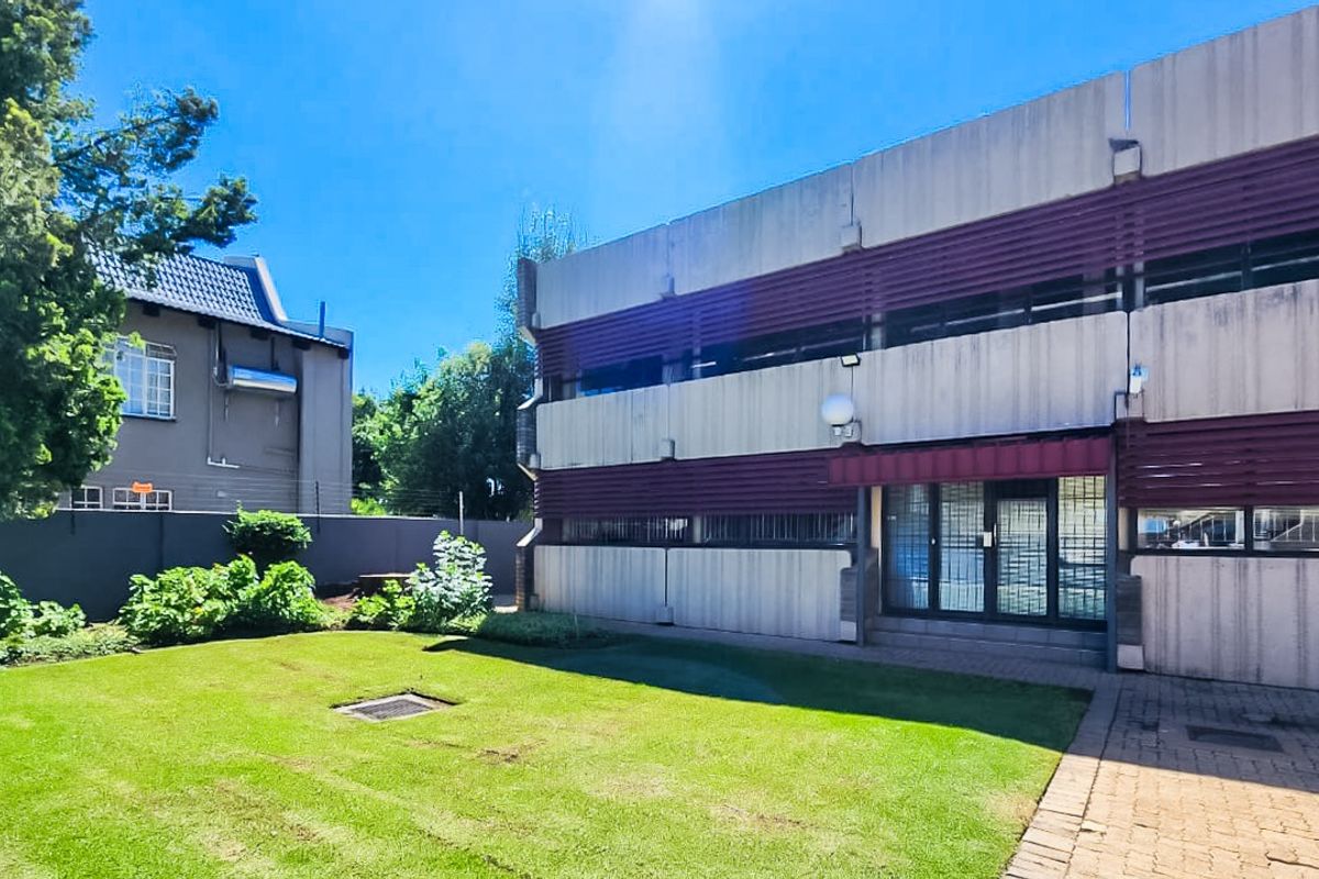 Rear view of a commercial building featuring a concrete facade with louvre shutters, a paved walkway, and a well-maintained lawn area.
