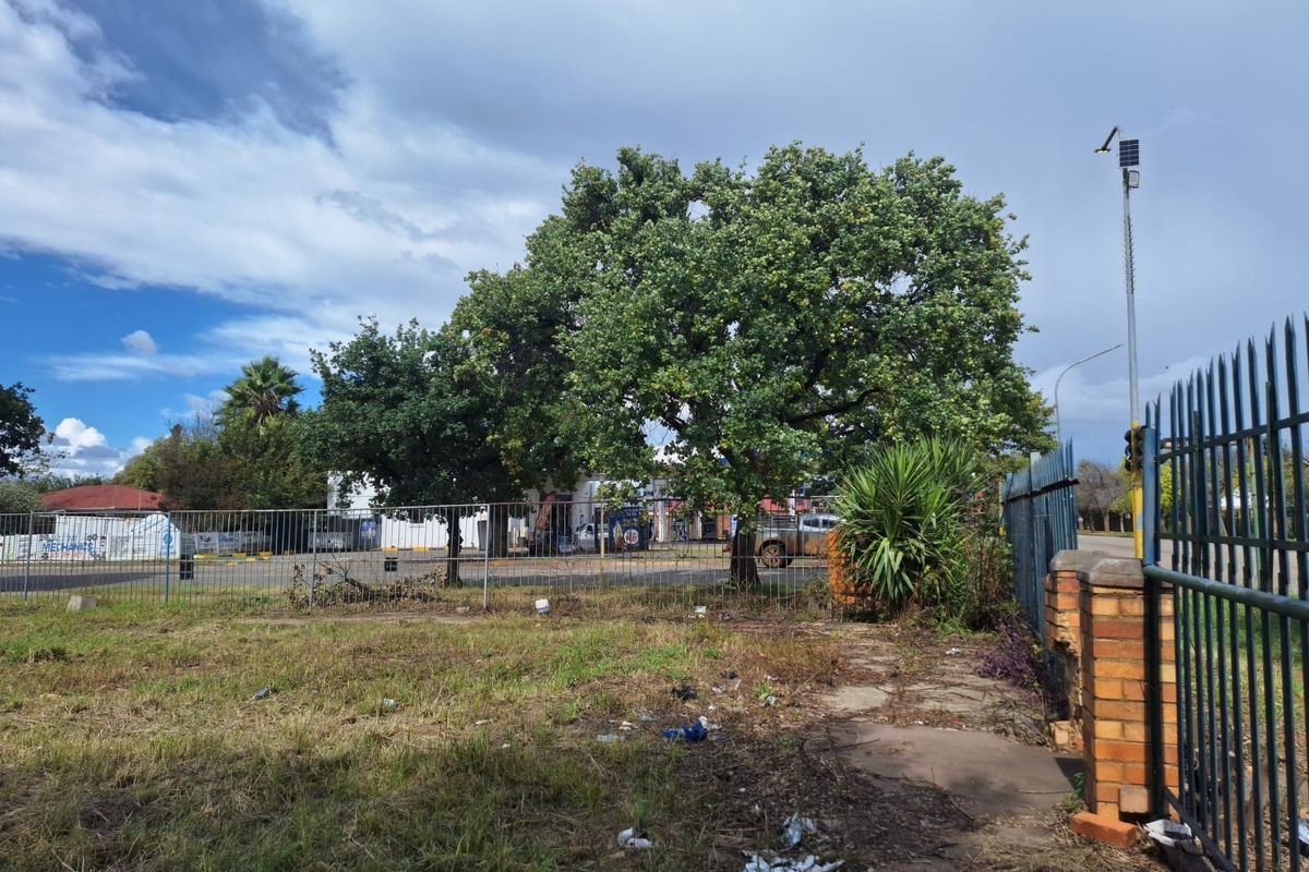 A vacant commercial plot featuring metal palisade fencing, a brick pillar, and mature trees, offering street-level access and visibility.