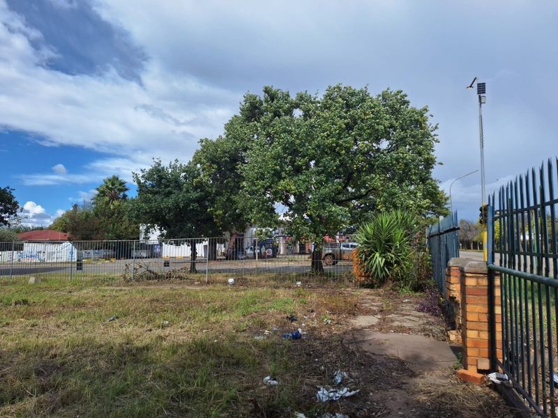 A vacant commercial plot featuring metal palisade fencing, a brick pillar, and mature trees, offering street-level access and visibility.