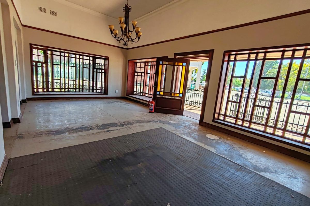 Spacious, empty living area featuring large timber-framed windows with security bars, a decorative chandelier, and a characterful stained glass entrance door.