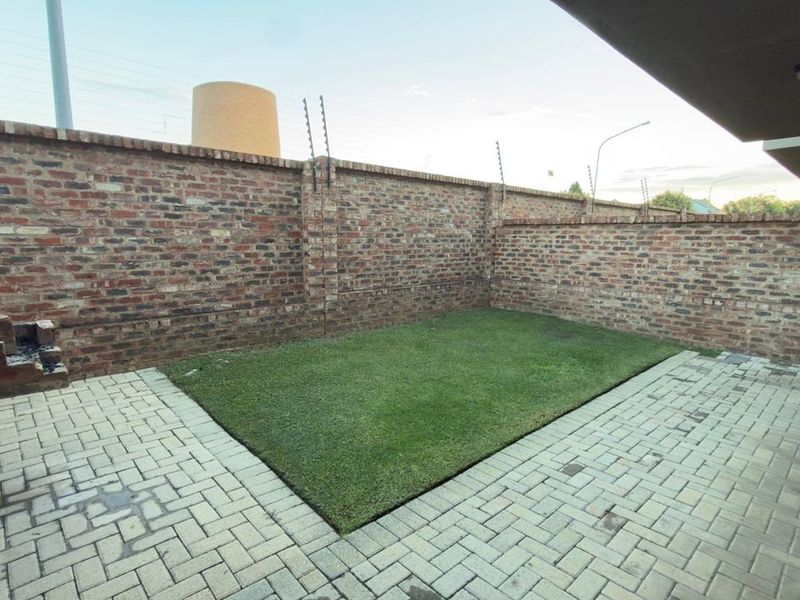 A private rear courtyard featuring a paved patio, artificial lawn, and a built-in brick braai, enclosed by high brick walls with electric fencing.