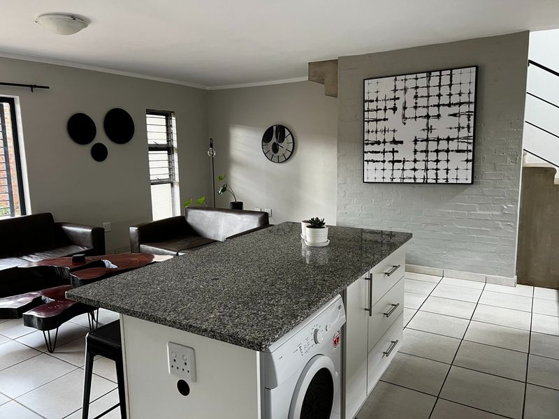 Open-plan living area featuring a kitchen island with granite countertop, tiled flooring, and a painted brick feature wall leading to the staircase.