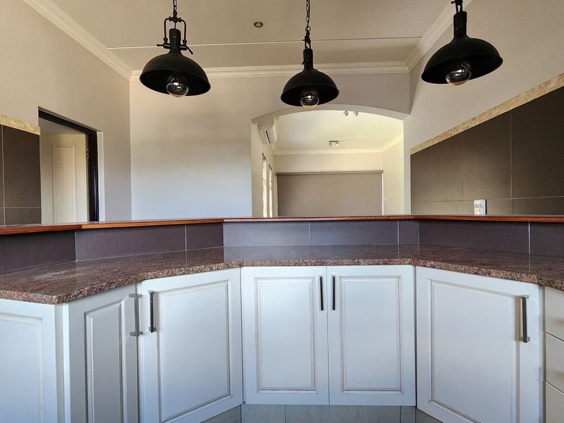 A clean kitchen area featuring white cabinetry, granite countertops, a breakfast bar, and modern pendant lighting, opening into an adjacent living space.