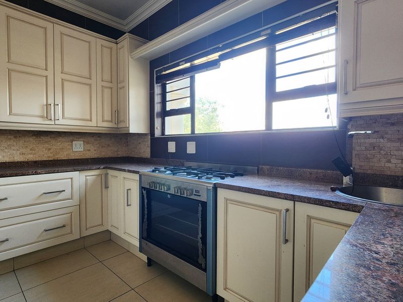 A functional kitchen featuring cream-colored cabinetry, stone countertops, a gas stove with oven, and a tiled backsplash with a large window.