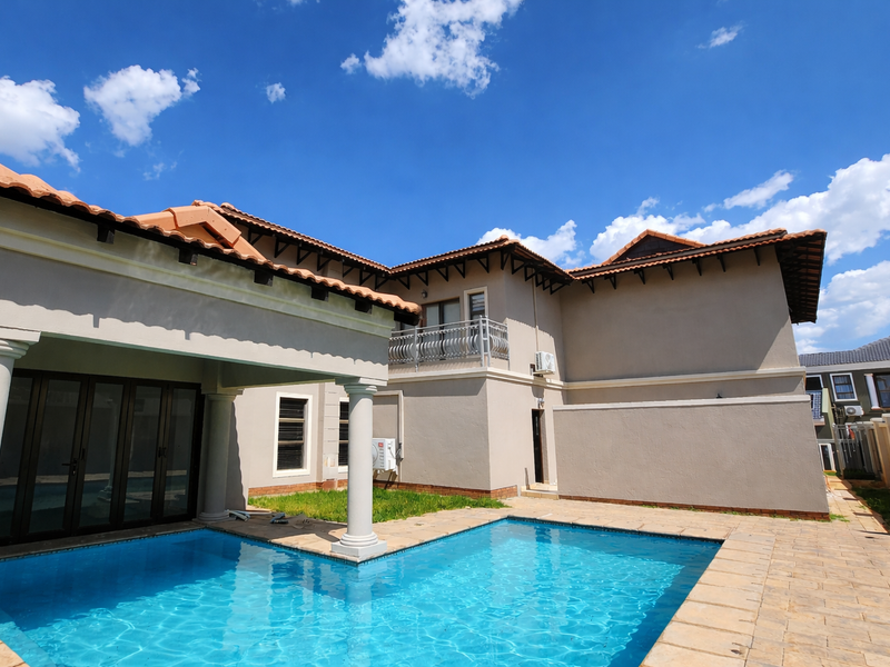 Rear view of the property featuring a swimming pool, covered patio area, and a balcony with decorative metal railing under a clear blue sky.