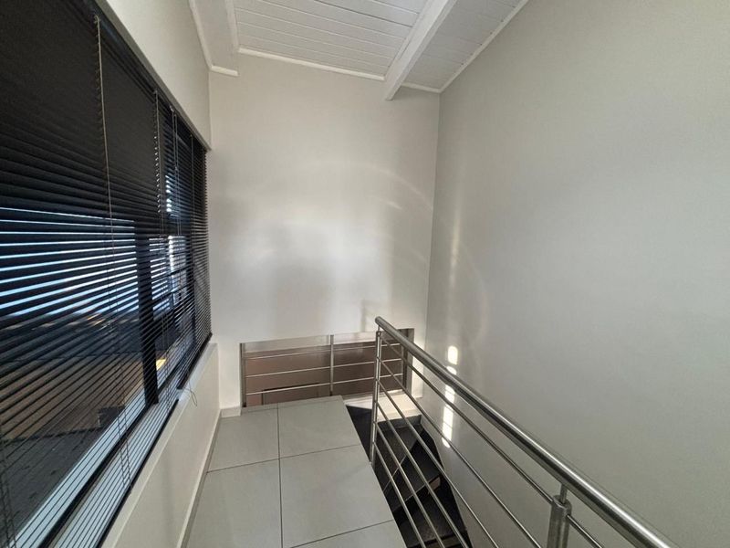 A bright, modern hallway featuring tiled flooring, a sleek stainless steel balustrade, and large windows fitted with venetian blinds.