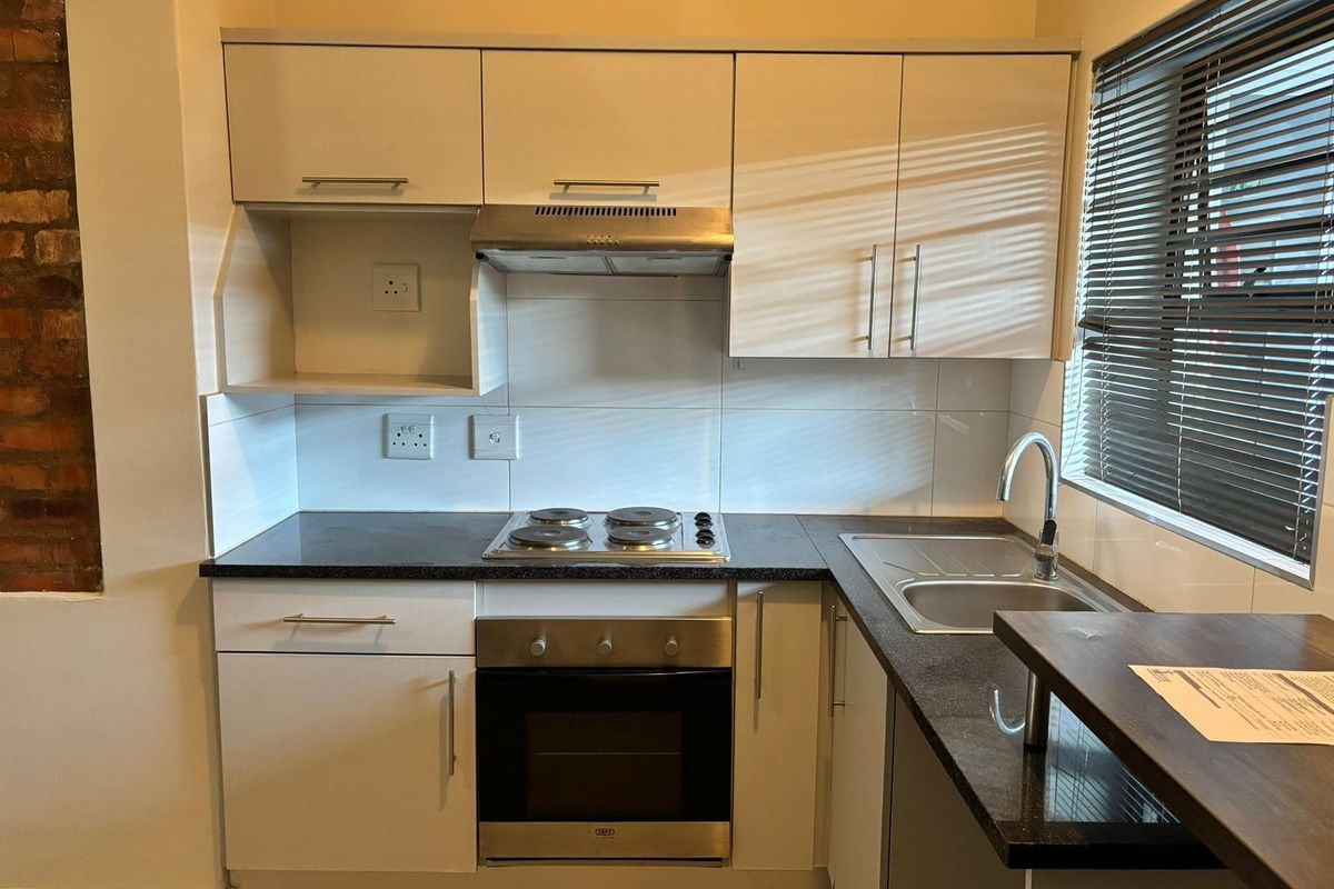 A compact, modern kitchen featuring white cabinetry, dark countertops, a built-in oven, electric hob, and a stainless steel sink with a tiled backsplash.
