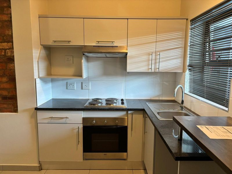 A compact, modern kitchen featuring white cabinetry, dark countertops, a built-in oven, electric hob, and a stainless steel sink with a tiled backsplash.