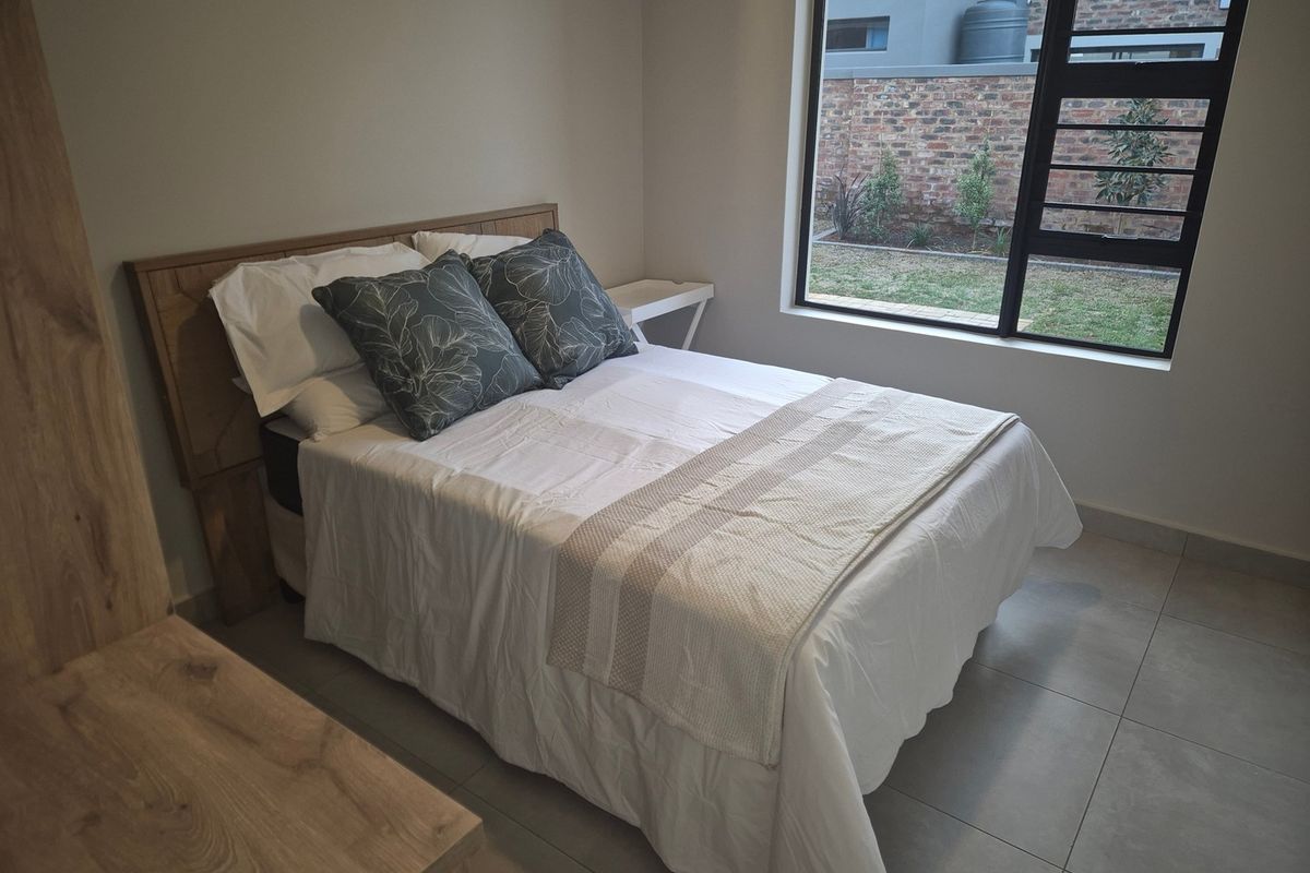 A well-lit bedroom featuring neutral tiled flooring, a large window with security bars, and a clean, modern aesthetic.