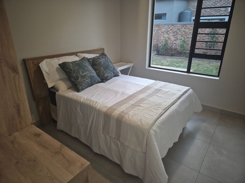 A well-lit bedroom featuring neutral tiled flooring, a large window with security bars, and a clean, modern aesthetic.