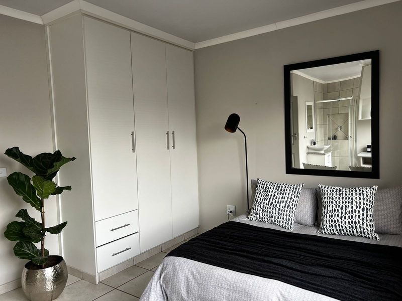 A well-maintained bedroom featuring neutral walls, tiled flooring, and ample built-in cupboard storage, with an adjoining en-suite bathroom visible through the mirror.