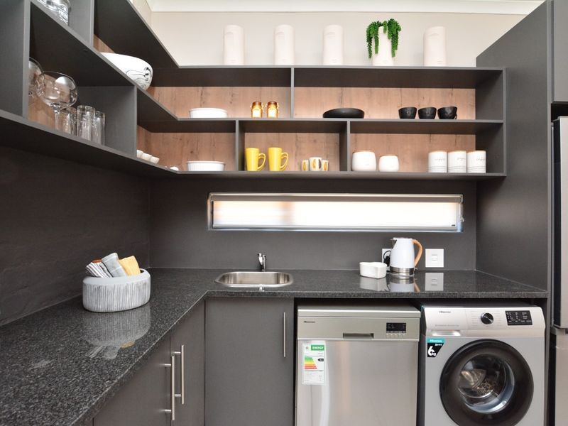 Modern scullery featuring dark cabinetry, granite countertops, integrated dishwasher, washing machine, and ample open shelving with integrated lighting.