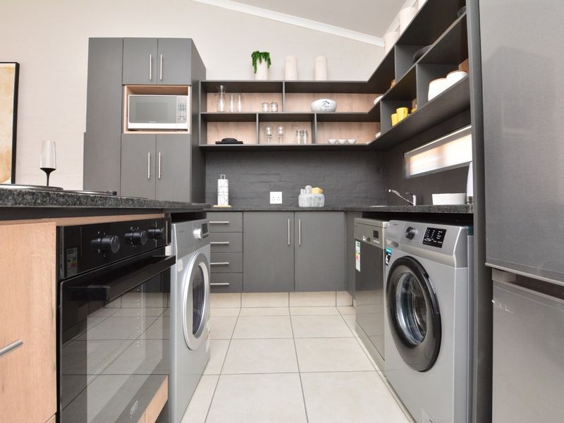 Modern scullery featuring dark grey cabinetry, granite countertops, built-in oven, microwave, and integrated laundry appliance connections with clean tiled flooring.