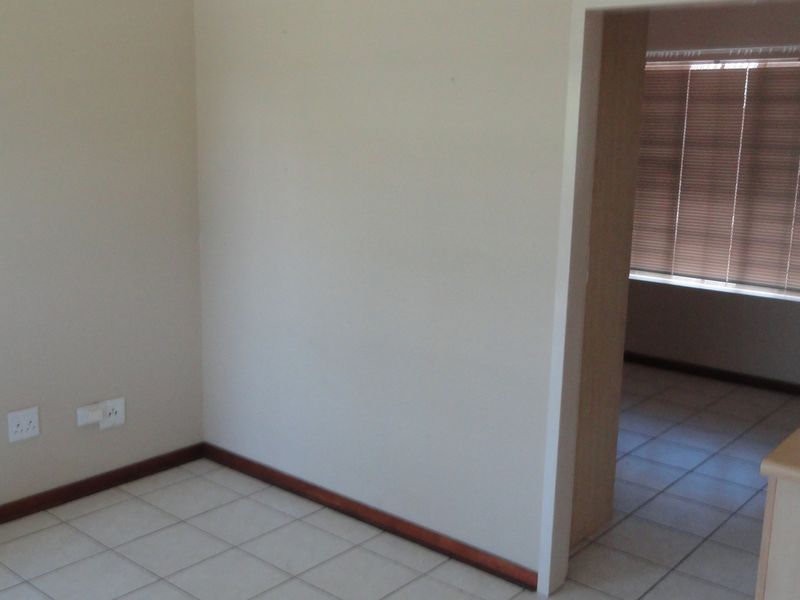 An empty open-plan living area featuring tiled flooring, neutral walls, and a view into an adjacent room with fitted venetian blinds.