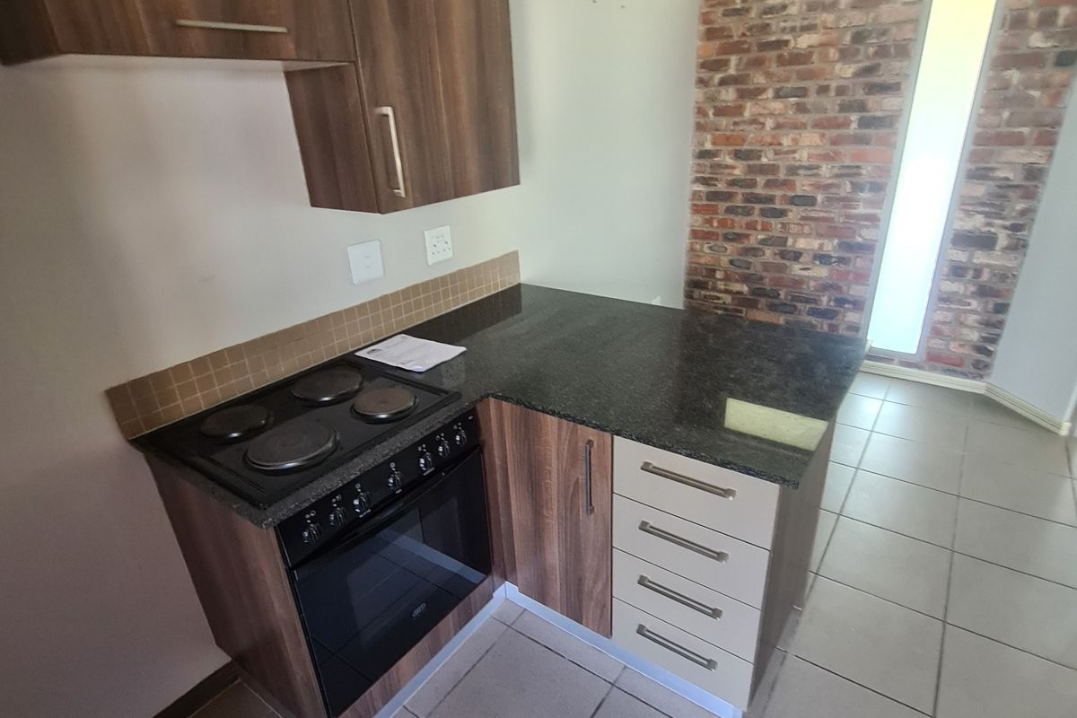 Compact kitchen featuring built-in cabinetry, granite countertops, an integrated electric stove and oven, tiled flooring, and a decorative exposed brick feature wall.
