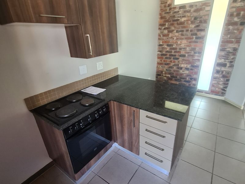 Compact kitchen featuring built-in cabinetry, granite countertops, an integrated electric stove and oven, tiled flooring, and a decorative exposed brick feature wall.