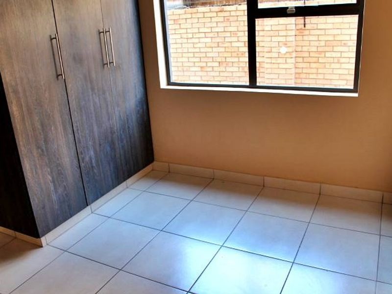 A clean, empty bedroom featuring modern dark wood-finish built-in cupboards, neutral tiled flooring, and a large window providing natural light.