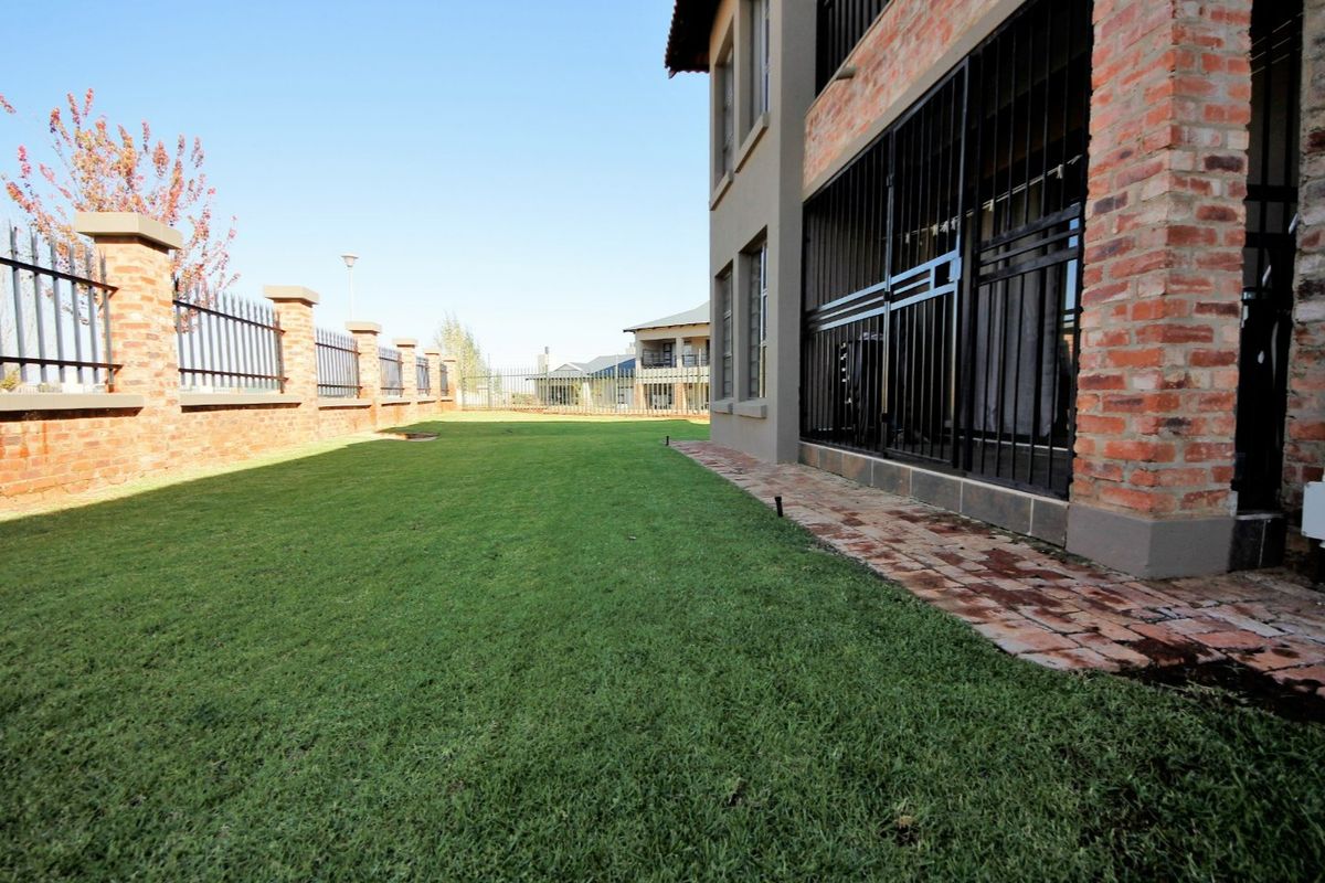 A well-maintained rear garden featuring a manicured lawn, brick boundary wall with security fencing, and a paved walkway along the building exterior.