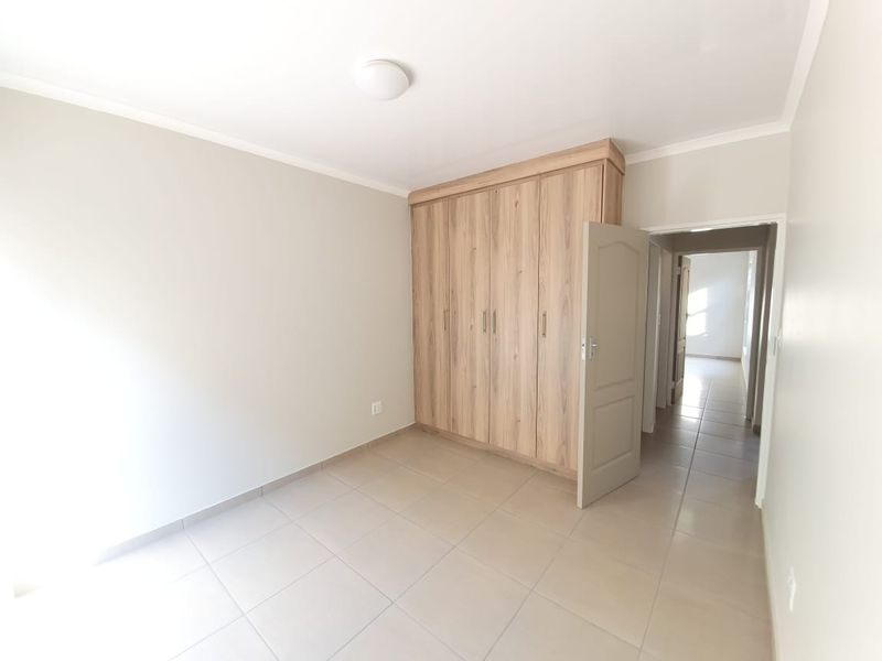 A bright, empty bedroom featuring neutral tiled flooring and a modern wood-finish built-in wardrobe, leading into a hallway.