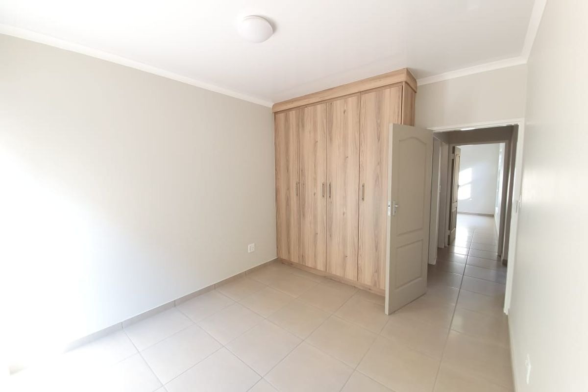 A bright, empty bedroom featuring neutral tiled flooring and a modern wood-finish built-in wardrobe, leading into a hallway.
