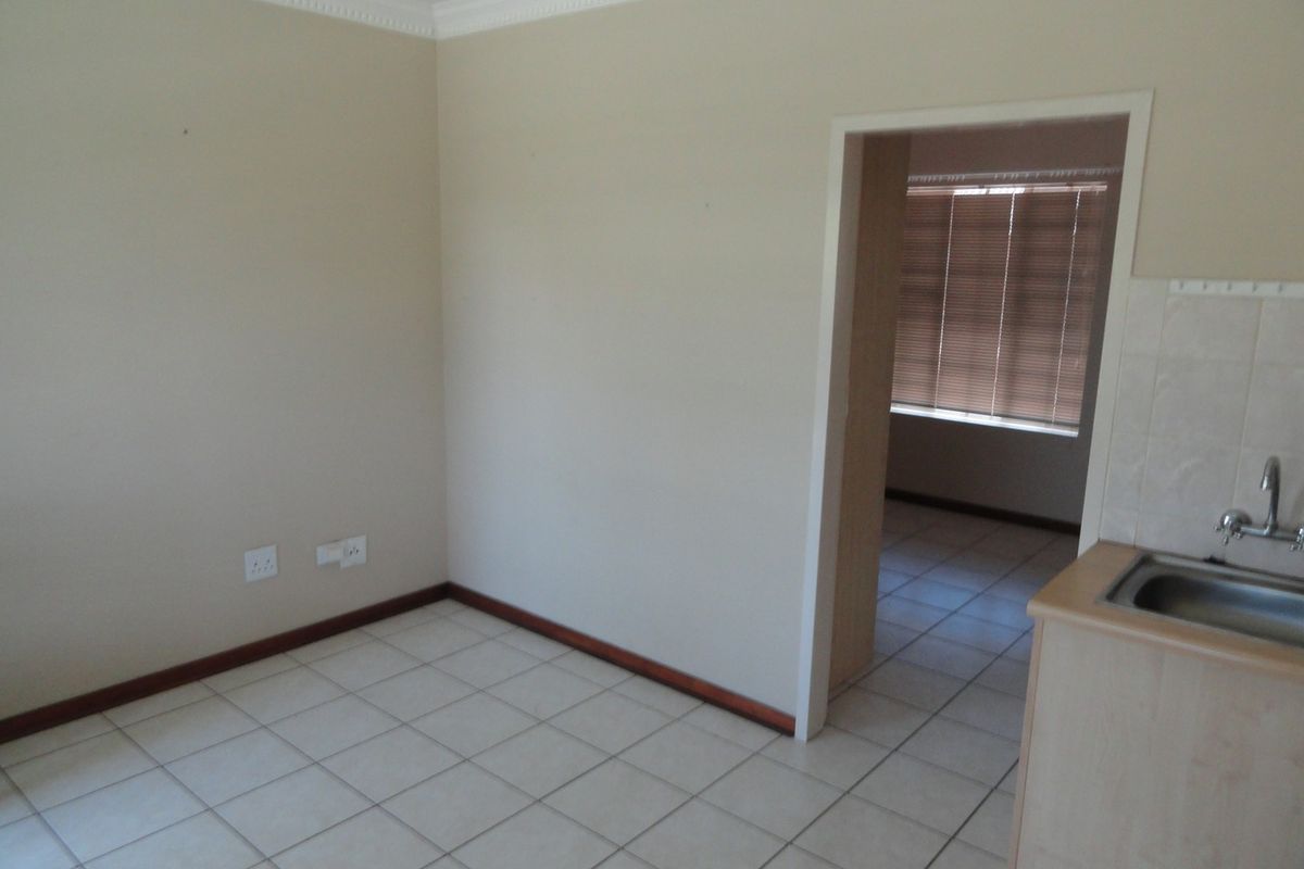 An open-plan living area featuring tiled flooring, crown molding, and a compact kitchen sink area, leading into an adjacent room with venetian blinds.