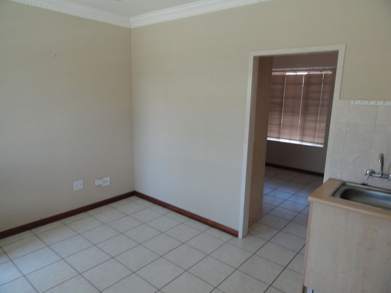 An open-plan living area featuring tiled flooring, crown molding, and a compact kitchen sink area, leading into an adjacent room with venetian blinds.