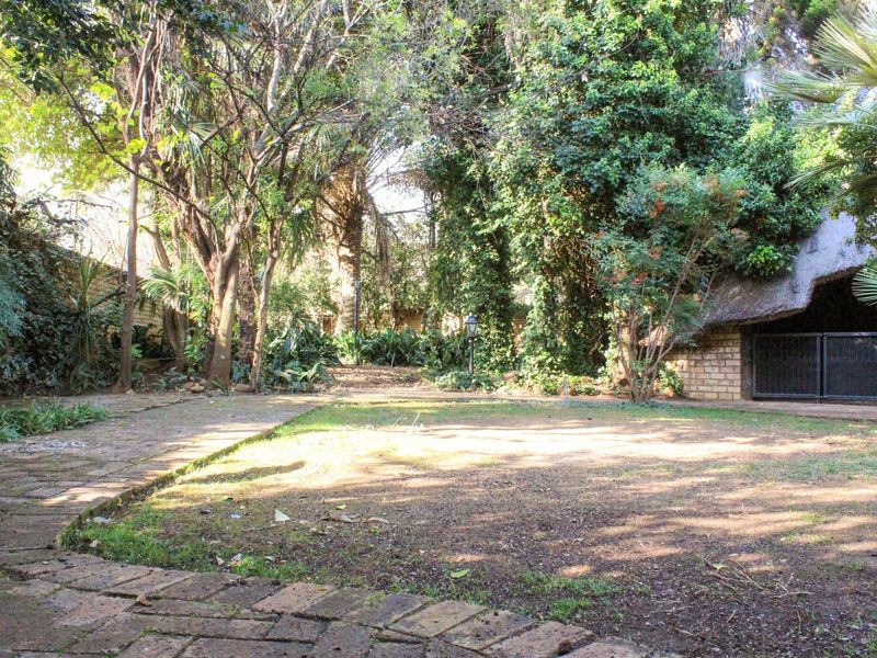 A private garden area featuring a brick-paved pathway, established trees, and a thatched-roof outdoor structure.