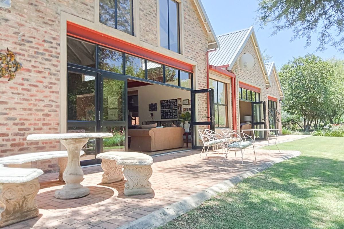 A charming rear exterior view of a brick farmhouse featuring expansive steel-framed folding doors that open onto a spacious paved patio and garden area.
