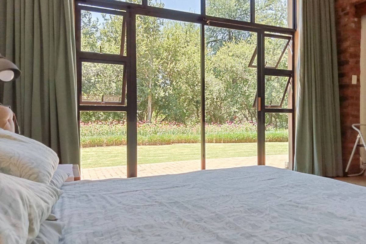 A bright bedroom featuring large steel-framed glass doors that open onto a lush garden, complemented by an exposed brick wall.