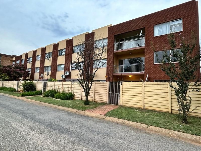 Exterior view of a multi-story apartment complex featuring red brick and cream facade, metal balcony railings, and a secure perimeter boundary wall.