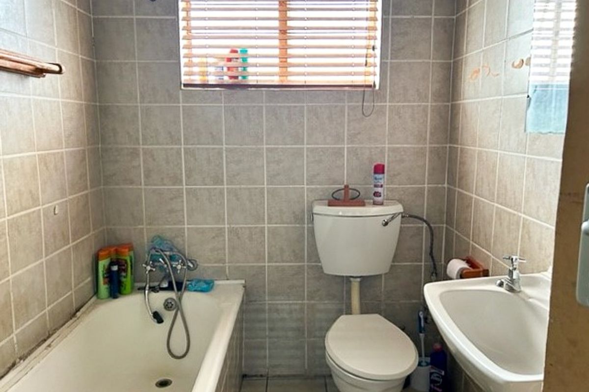 A functional bathroom featuring a standard bathtub, pedestal basin, and toilet, finished with neutral wall tiles and a window with fitted blinds.